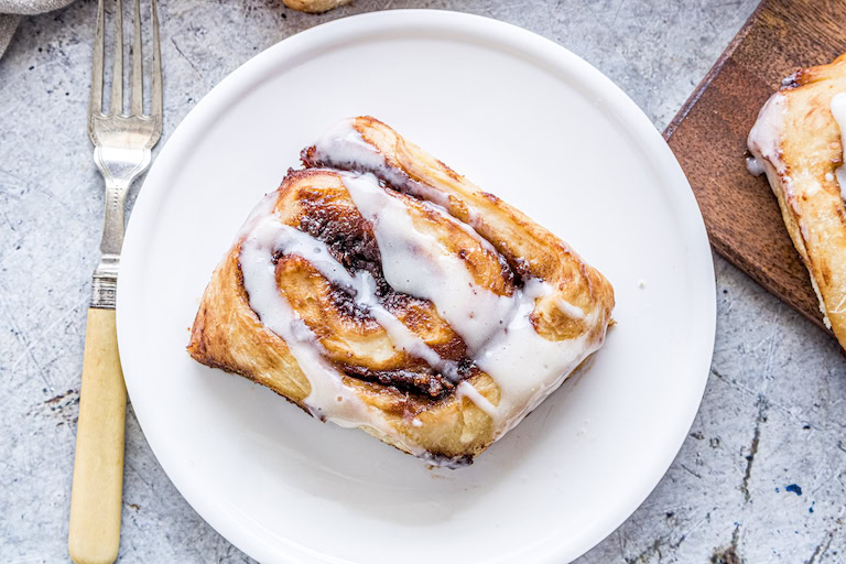 A cinnamon roll with white icing on a white plate, next to a fork on a light-colored surface.