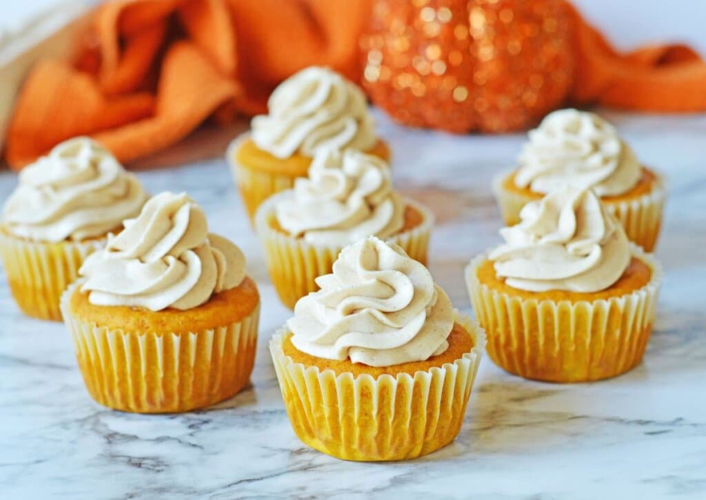 Seven pumpkin cupcakes with swirled cream frosting are arranged on a marble surface, with an orange cloth and pumpkin decoration in the background.