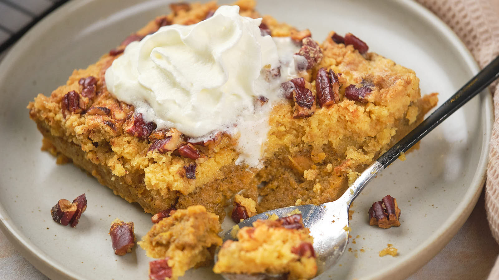 A slice of pumpkin dessert topped with whipped cream and chopped pecans on a plate with a spoon.