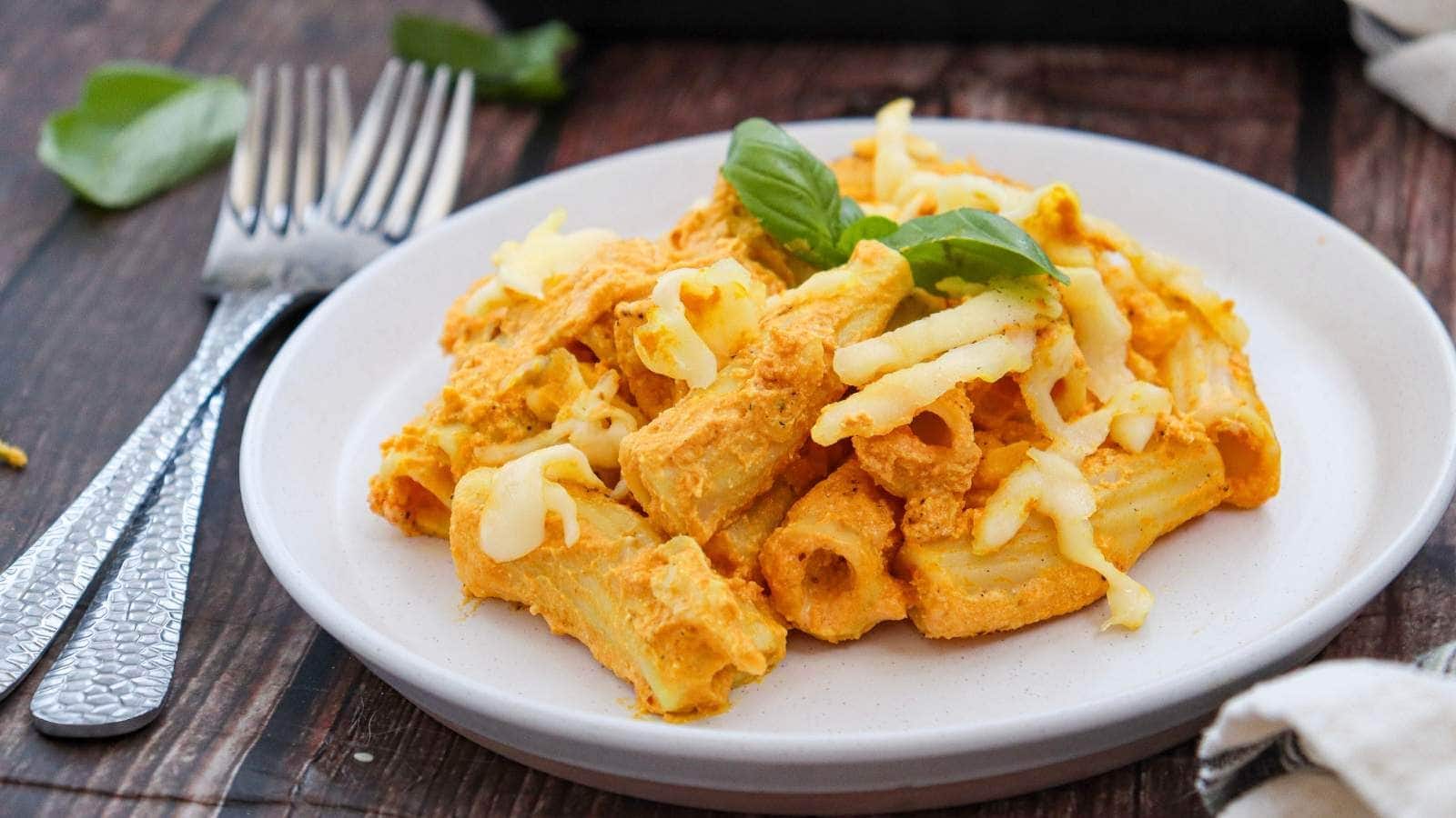 A plate of rigatoni pasta with creamy orange sauce, topped with melted cheese and garnished with a basil leaf, placed next to two forks on a wooden table.