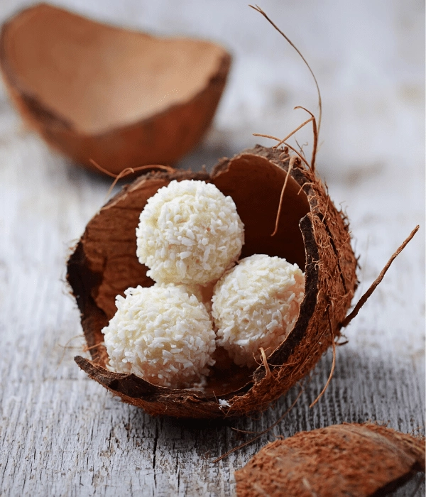 Three white coconut balls sit inside a halved coconut shell on a rustic wooden surface, with bits of coconut shell nearby.