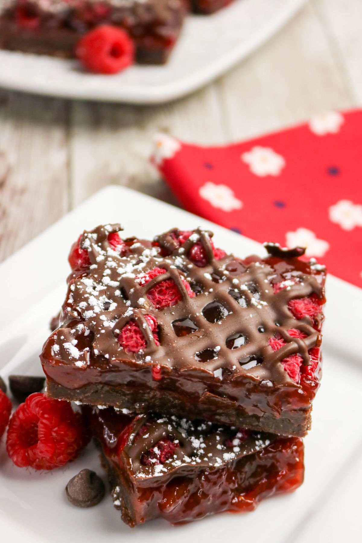 Two chocolate raspberry bars topped with powdered sugar and chocolate drizzle are stacked on a white plate, with a red napkin and scattered raspberries nearby.