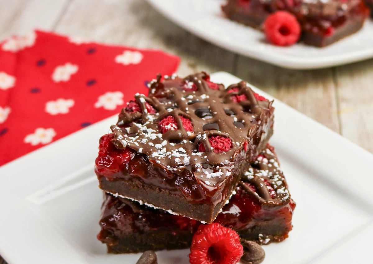 Two dessert bars topped with raspberries, chocolate drizzle, and powdered sugar are stacked on a white plate, with a red napkin and another plate in the background.