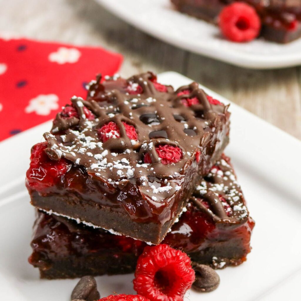 Two chocolate raspberry brownies topped with chocolate drizzle, powdered sugar, and fresh raspberries are stacked on a white plate with a few chocolate chips and raspberries beside them.