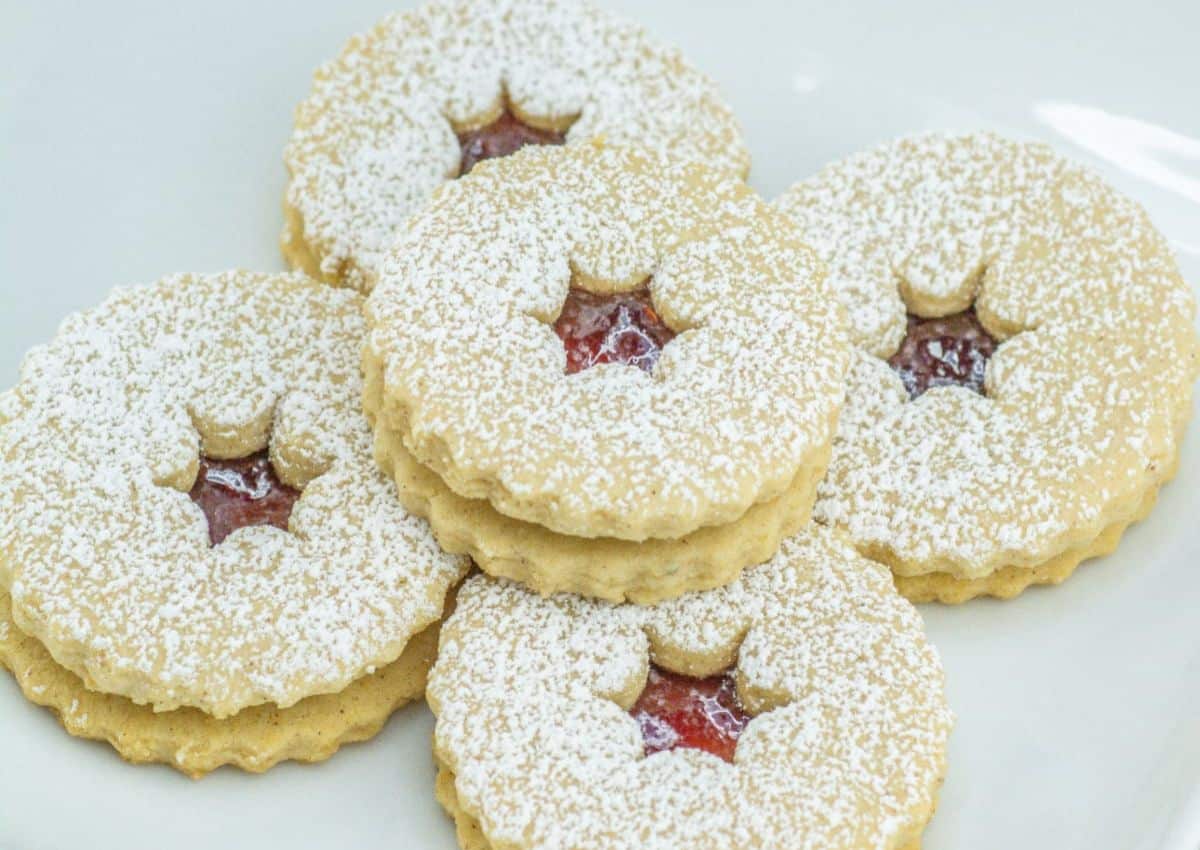 Five round sandwich cookies with scalloped edges, filled with red jam and dusted with powdered sugar, are arranged on a white surface.