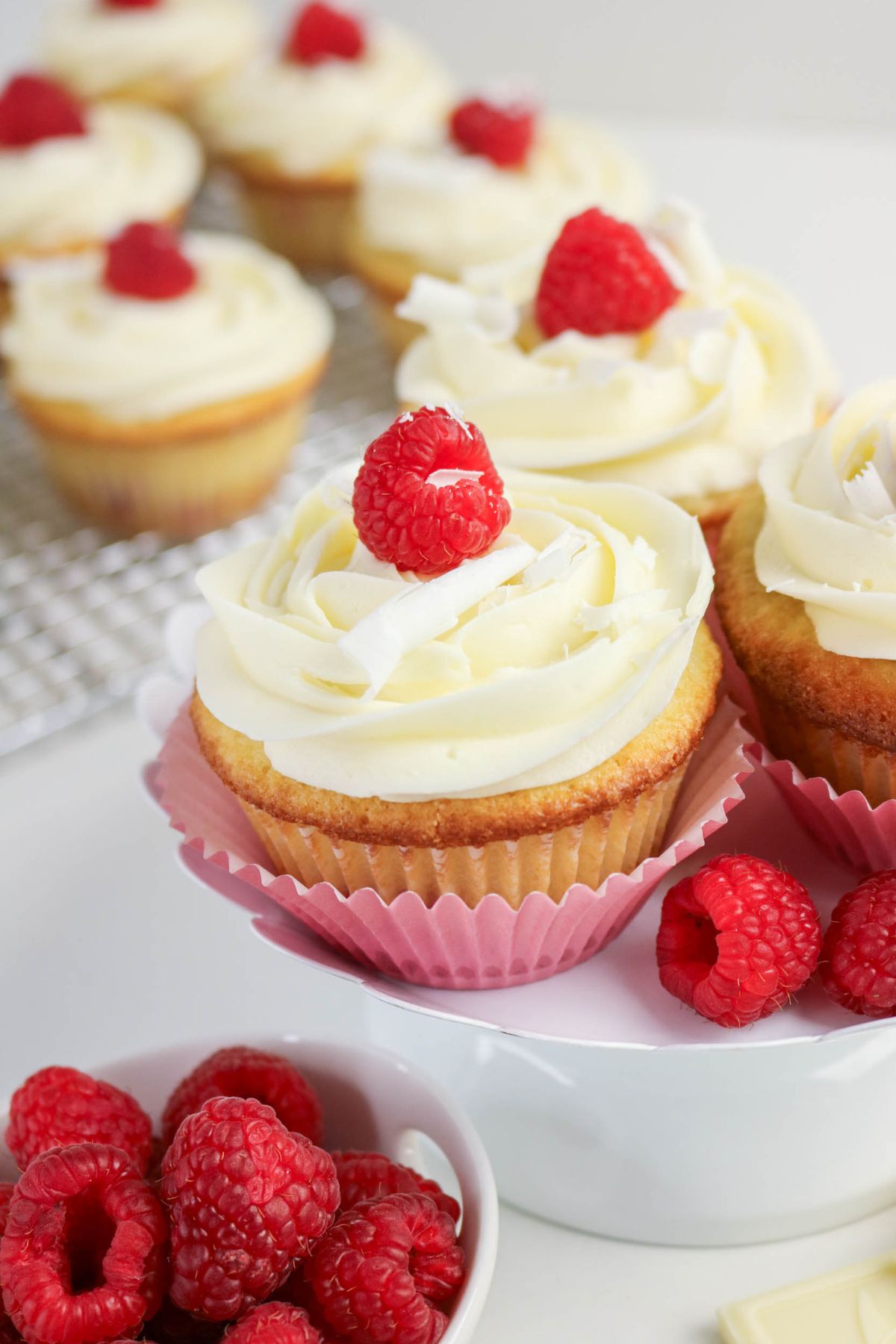 Cupcakes with white frosting and raspberries on top are displayed on a stand, with fresh raspberries in a bowl nearby and more cupcakes on a cooling rack in the background.