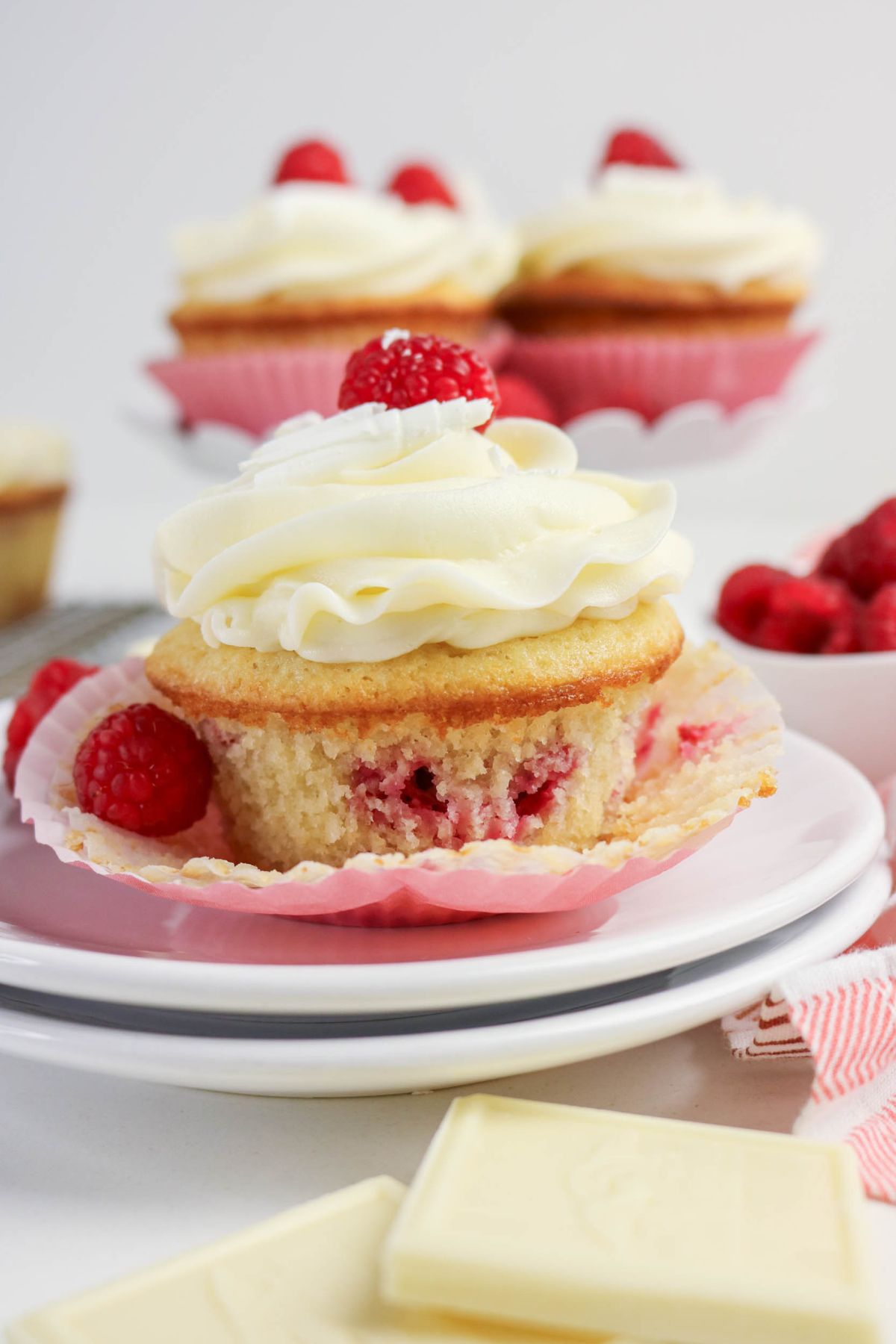 A raspberry cupcake with white frosting and a raspberry on top sits on a plate, surrounded by fresh raspberries and pieces of white chocolate.