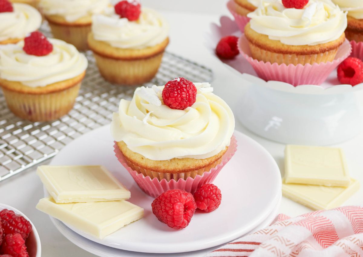 Vanilla cupcakes with white frosting are topped with raspberries and served with pieces of white chocolate on a white plate. Additional cupcakes and raspberries are in the background.
