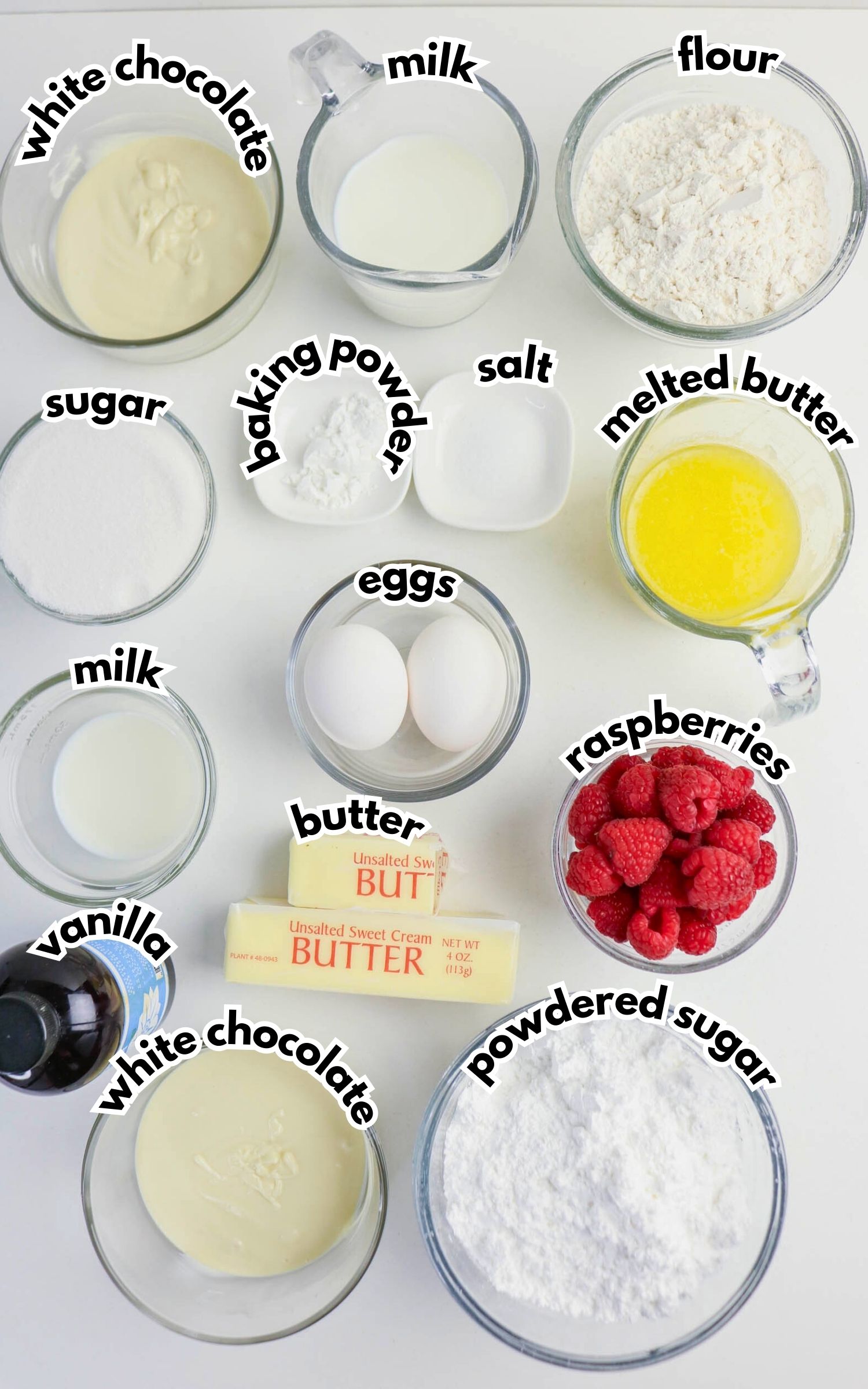 Top-down view of labeled baking ingredients in bowls and cups, including white chocolate, flour, sugar, baking powder, salt, melted butter, eggs, raspberries, vanilla, and powdered sugar.