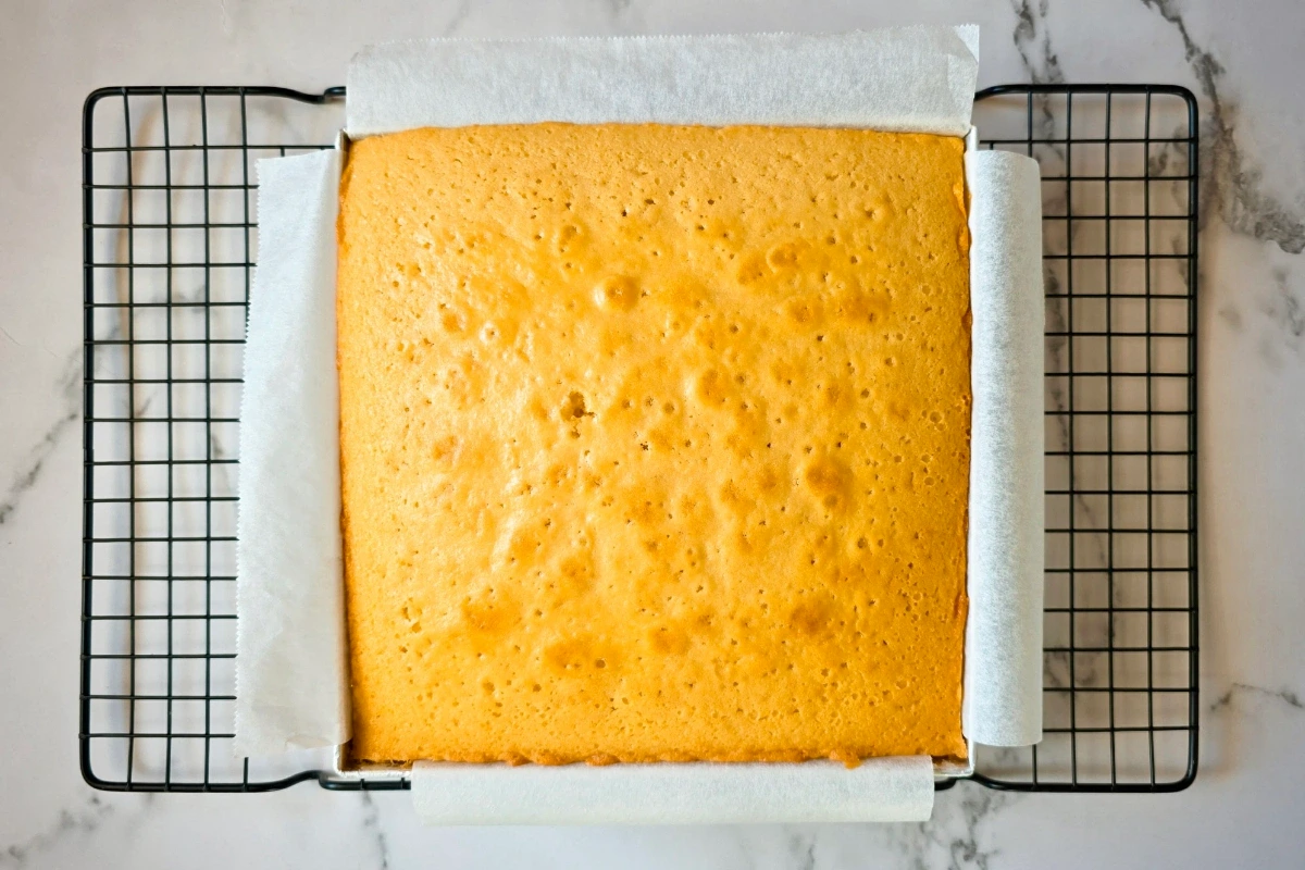 A square sponge cake in a baking tray lined with parchment paper, cooling on a wire rack on a marble surface.