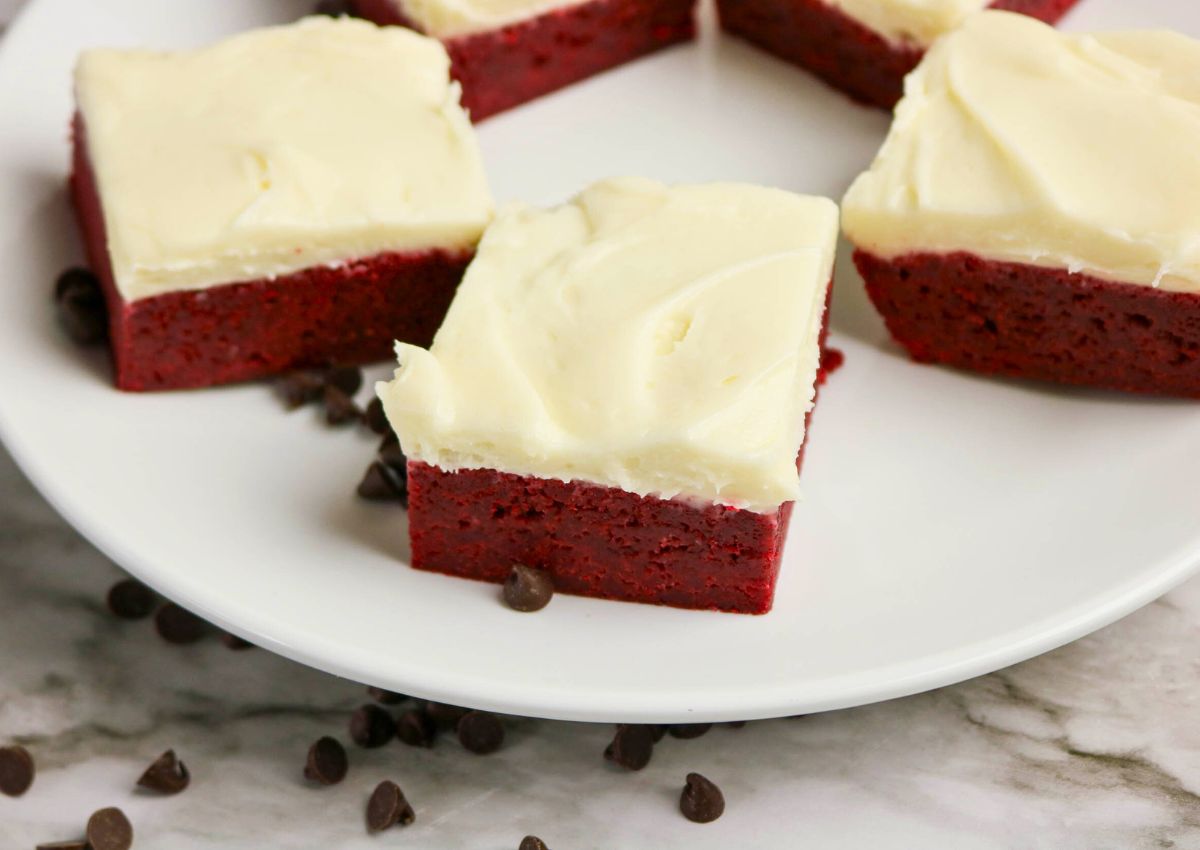 Squares of red velvet brownies with a layer of white frosting are arranged on a white plate, with chocolate chips scattered nearby.