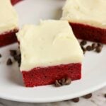 A square piece of red velvet cake with cream cheese frosting on a white plate, surrounded by a few chocolate chips.