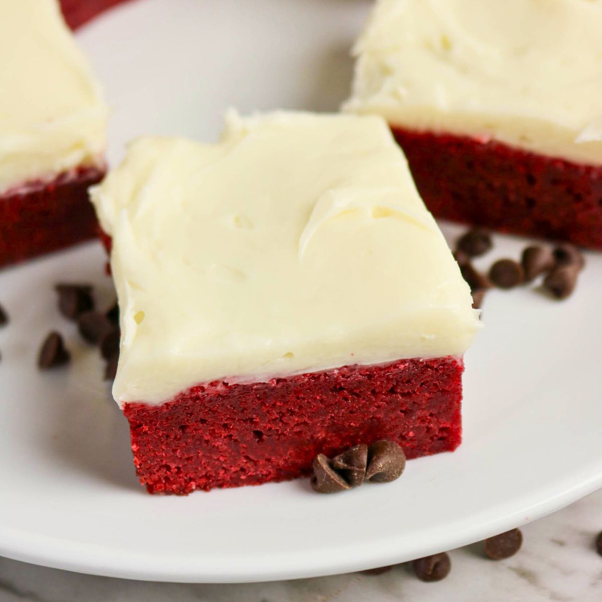 A square piece of red velvet cake with cream cheese frosting on a white plate, surrounded by a few chocolate chips.