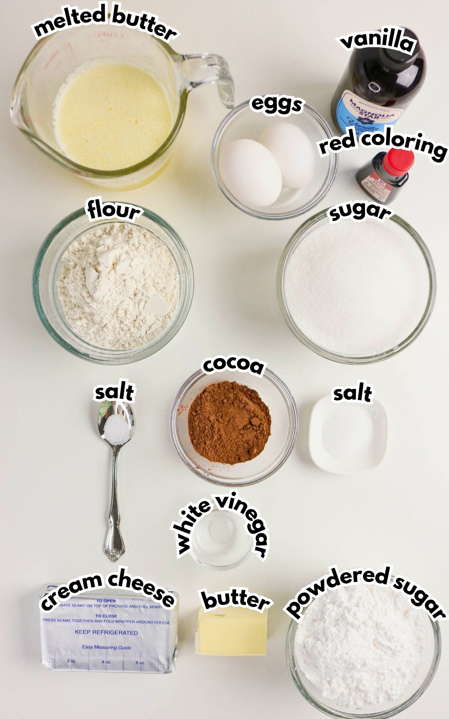 Various baking ingredients are arranged on a white surface, including melted butter, eggs, vanilla, red coloring, flour, sugar, cocoa, salt, cream cheese, butter, white vinegar, and powdered sugar.