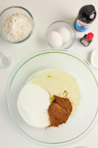 A glass bowl with sugar, cocoa powder, and melted butter, surrounded by bowls of flour, eggs, and bottles of vanilla and red food coloring.