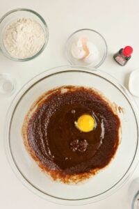 A glass bowl with brown batter and a raw egg, surrounded by bowls of flour, eggshells, vanilla extract, and a small dish on a white surface.