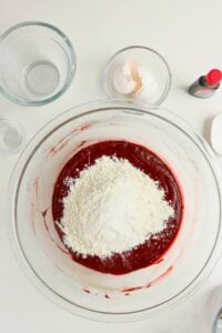 A large glass bowl with red batter and flour on top, surrounded by empty glass bowls, eggshells, and a bottle of red food coloring on a white surface.