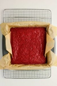A square red velvet cake in a parchment-lined baking pan, cooling on a wire rack against a white background.