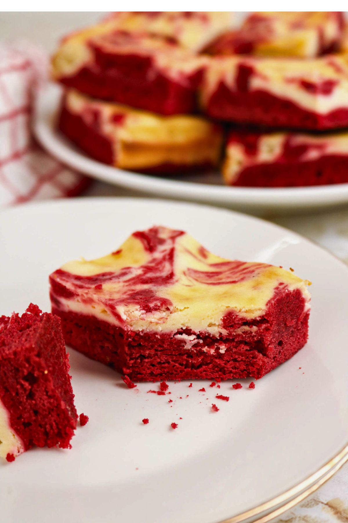 A partially eaten red velvet cheesecake brownie sits on a white plate, with more brownies stacked on a plate in the background.