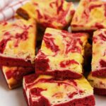Red velvet cheesecake brownies stacked on a plate, showing marbled red and cream-colored layers.