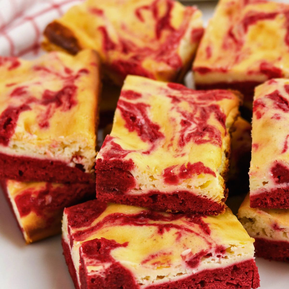 Red velvet cheesecake brownies stacked on a plate, showing marbled red and cream-colored layers.