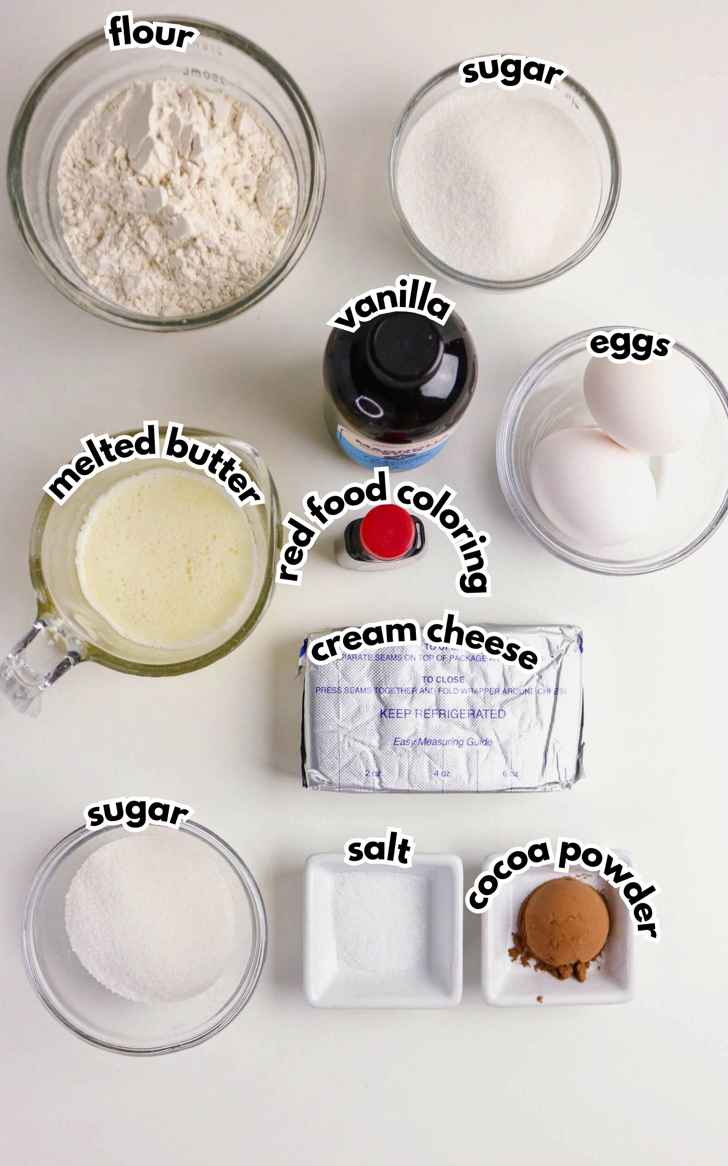 Baking ingredients arranged on a white surface: flour, sugar, vanilla, eggs, melted butter, red food coloring, cream cheese, salt, and cocoa powder, each labeled.