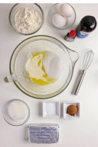 Ingredients for baking, including flour, eggs, sugar, vanilla extract, whisk, baking soda, cocoa powder, butter, and salt arranged on a white surface.