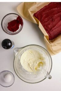 Overhead view of baking ingredients: a pan of red cake, a bowl of whipped cream cheese frosting, an egg, red batter, and a bottle of vanilla extract on a white surface.