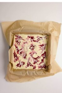 A square baking pan lined with parchment paper holds unbaked cheesecake batter swirled with red raspberry sauce.