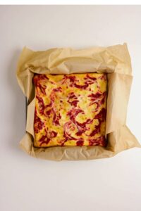 A square cheesecake with red fruit swirls on top, baked in a parchment-lined pan.
