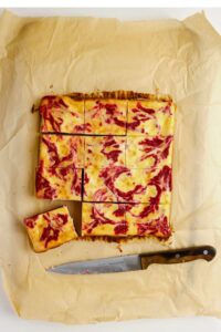Nine cheesecake bars with a raspberry swirl pattern are cut and arranged on parchment paper, with a knife placed beside them.