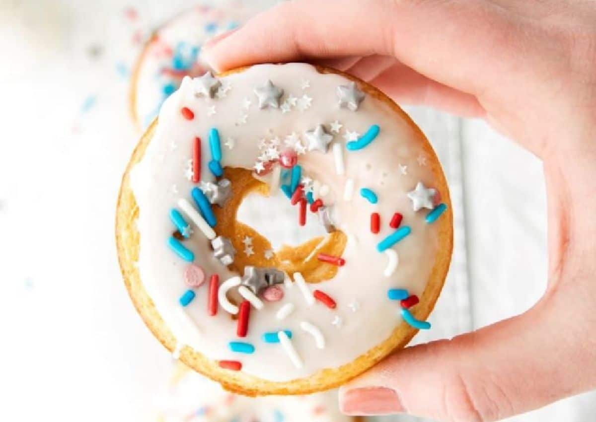A hand holds a donut topped with white icing and red, white, blue, and silver star-shaped sprinkles.