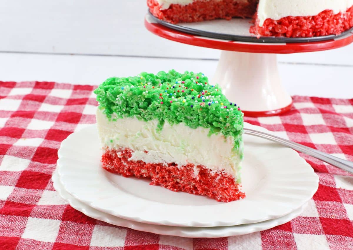 A slice of layered dessert with red and green cake, white cream filling, and colorful sprinkles on top, served on a white plate with a fork.