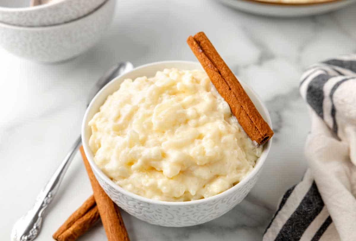 A bowl of rice pudding garnished with two cinnamon sticks, placed on a marble surface next to a striped napkin and a spoon.