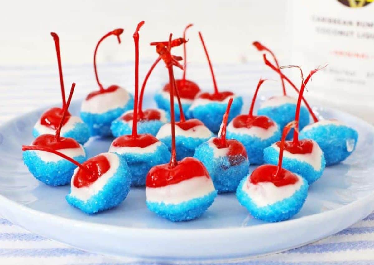 A plate of cherries dipped in white coating and blue sugar, arranged on a white plate with visible stems.