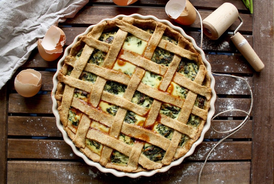 A baked savory pie with a lattice crust sits on a wooden surface, surrounded by cracked eggshells, a rolling pin, flour, and a cloth.
