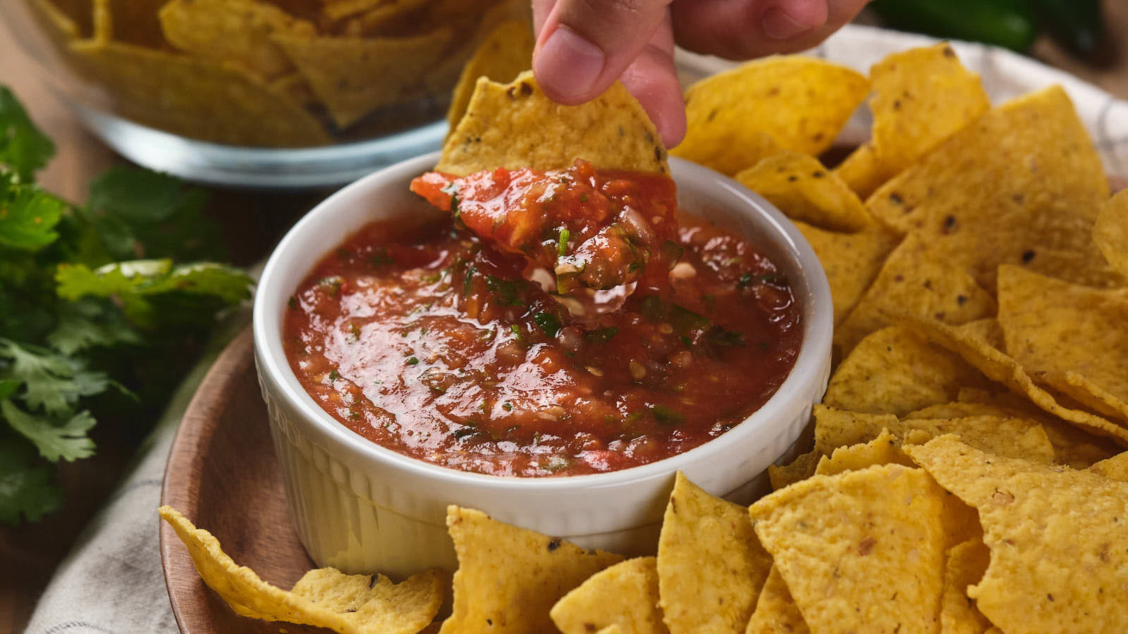 A hand dips a tortilla chip into a bowl of salsa, surrounded by more tortilla chips on a wooden plate.