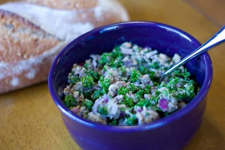 A purple bowl filled with a salad containing chopped broccoli, tuna, red onion, and herbs, with a spoon inside. A loaf of bread sits in the background on a wooden surface.