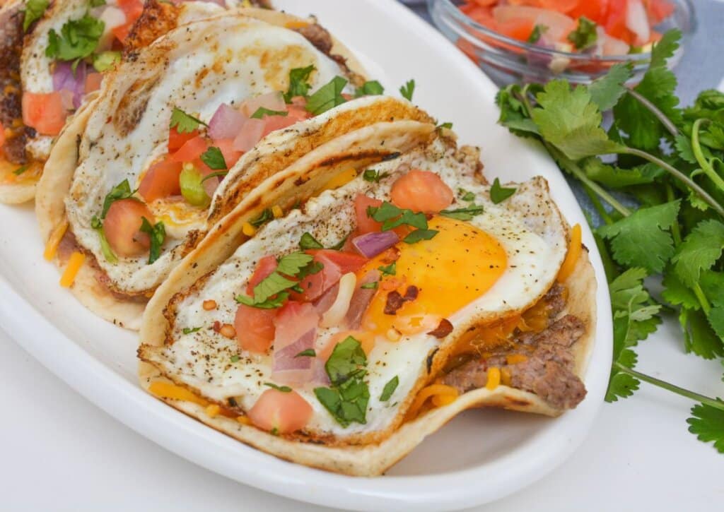 Three breakfast tacos filled with refried beans, cheddar cheese, fried eggs, diced tomatoes, onions, and fresh cilantro are served on a white plate with cilantro garnish on the side.