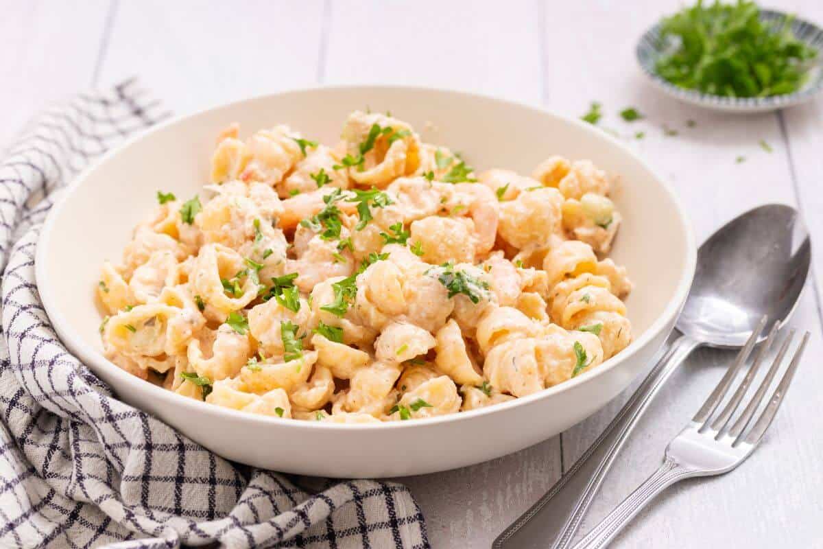 A white bowl of creamy shell pasta garnished with chopped parsley, placed on a striped cloth with a spoon and fork beside it.