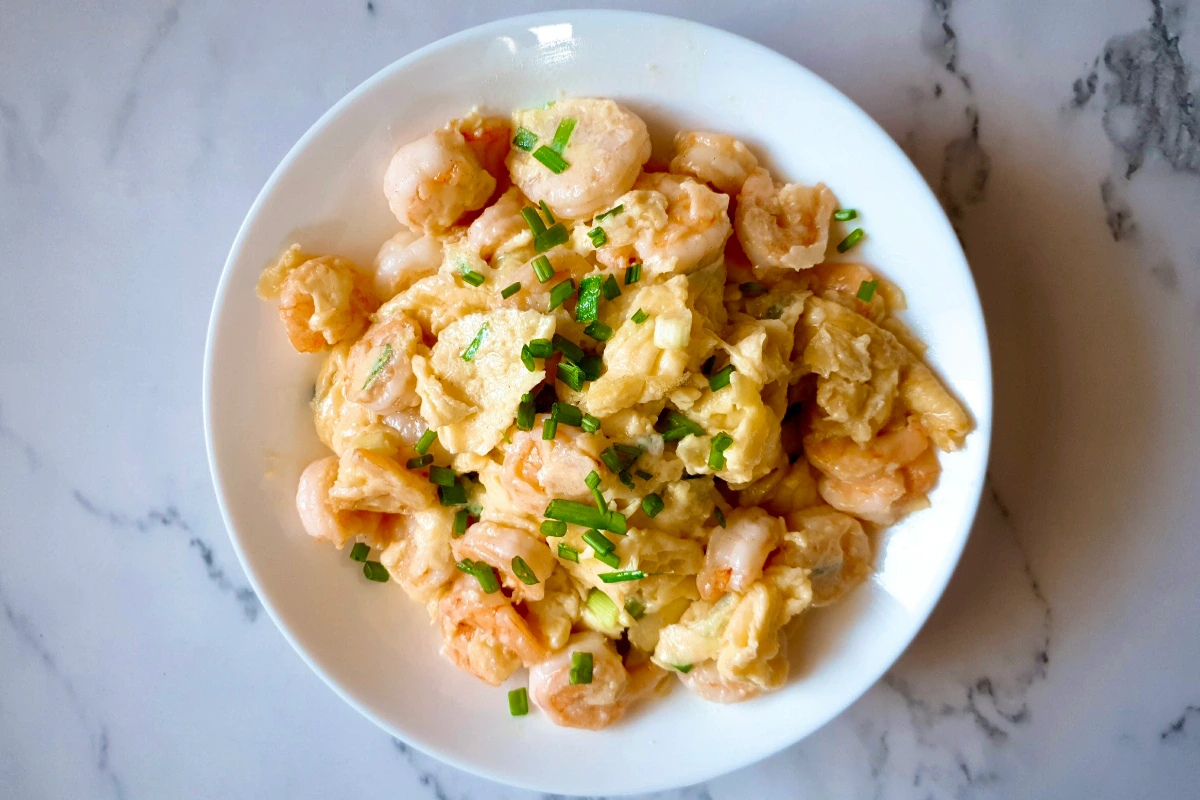A white plate with scrambled eggs and shrimp, garnished with chopped chives, on a marble countertop.