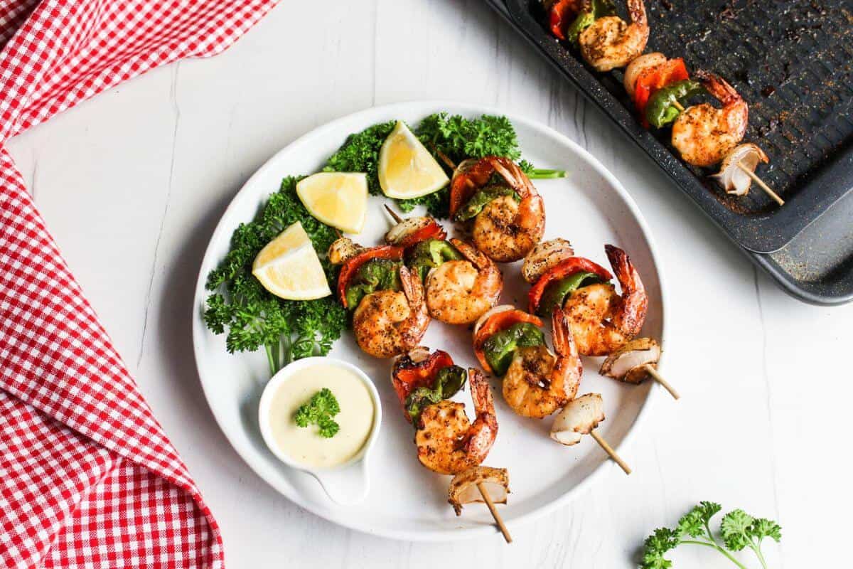 Grilled shrimp and vegetable skewers served on a white plate with lemon wedges, parsley, and a side of dipping sauce; a grill pan and red checkered cloth are nearby.