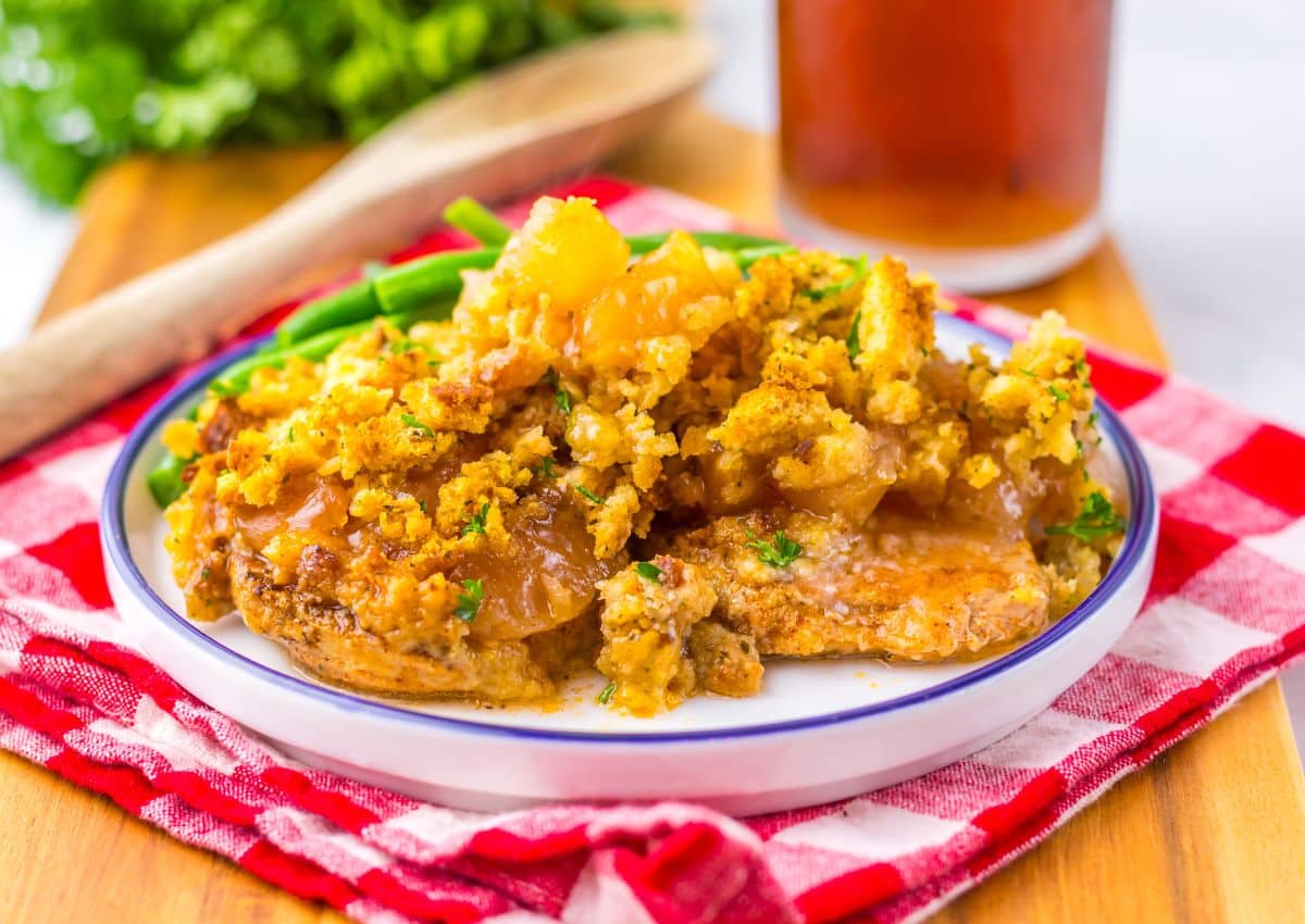 A plate of baked pork chops topped with a golden cornbread stuffing sits on a red plaid napkin, with green beans and a drink in the background.