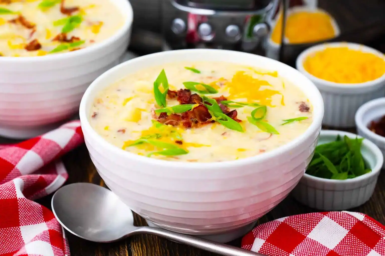 A bowl of creamy potato soup topped with chopped green onions, shredded cheese, and bacon, with a spoon and condiments on the side.