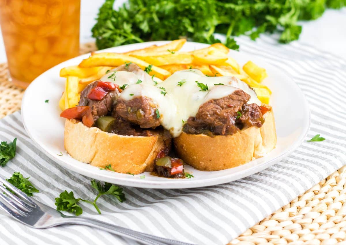Open-faced sandwiches topped with beef, melted cheese, and diced peppers, served with French fries on a white plate, garnished with parsley.