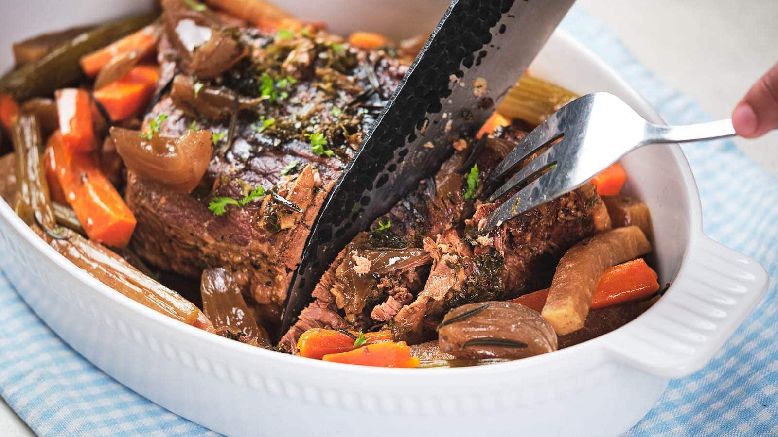 A cooked pot roast with carrots, onions, and celery in a white baking dish is being sliced with a knife and held with a fork.