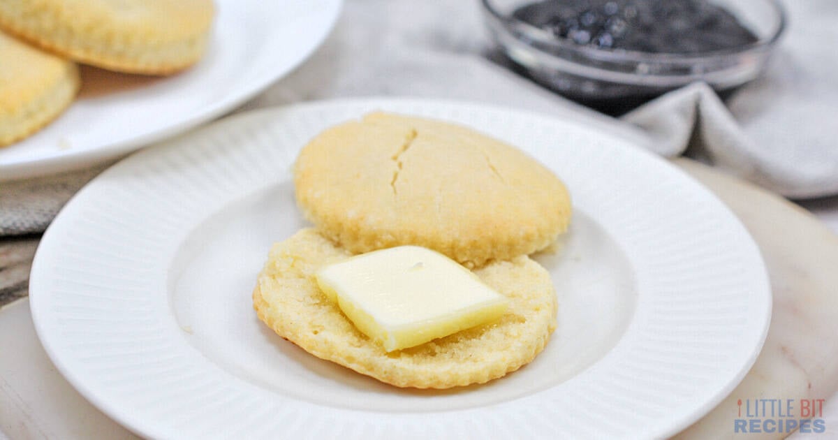 A split biscuit on a white plate with a pat of butter on one half; a plate of more biscuits and a bowl of jam are in the background.
