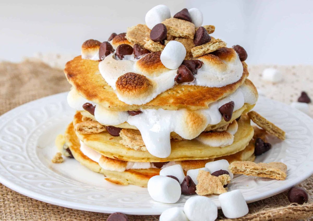 A stack of pancakes topped with toasted marshmallows, chocolate chips, and graham cracker pieces on a white plate.