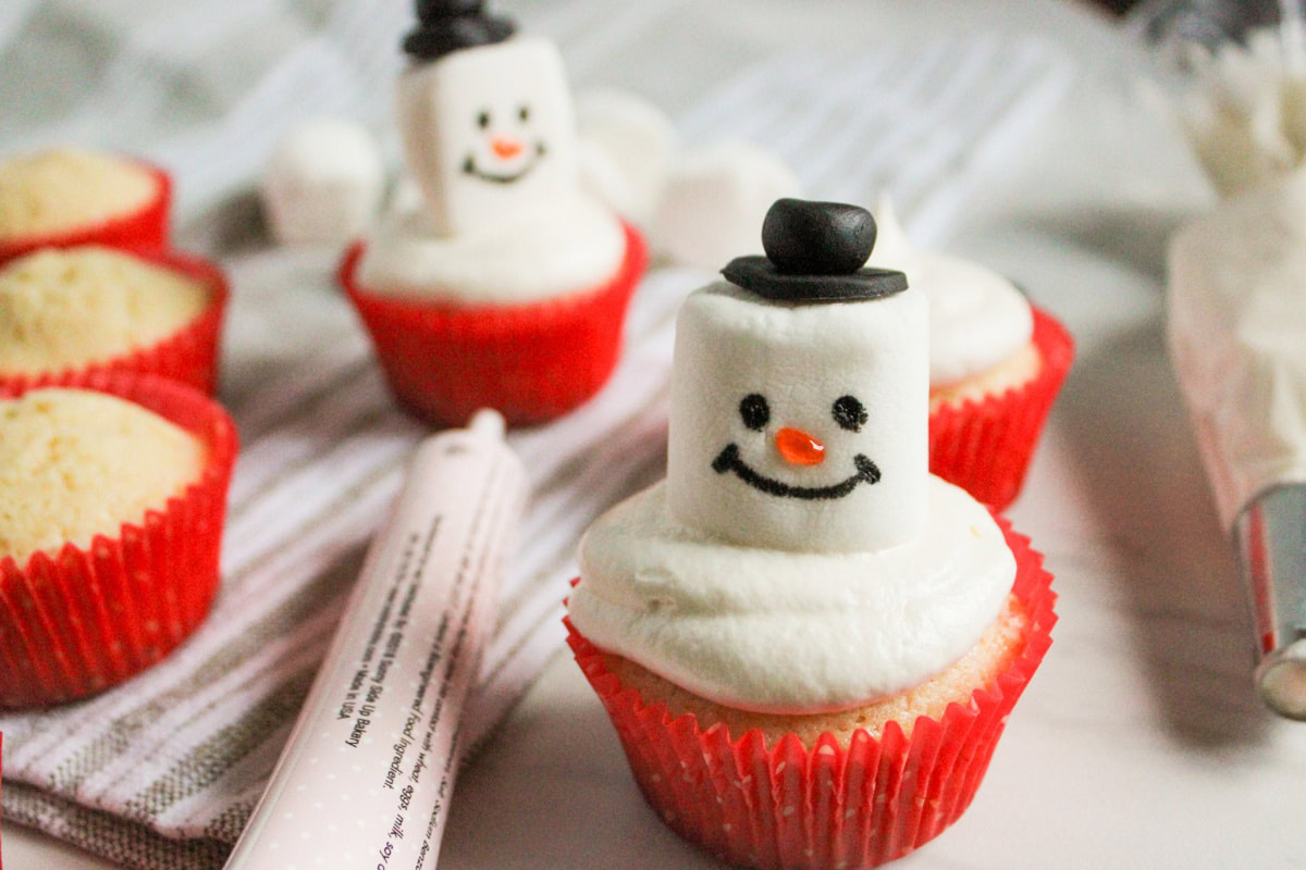 Cupcakes with white icing, topped with marshmallows decorated as snowmen, in red paper liners on a light surface with decorating tools nearby.
