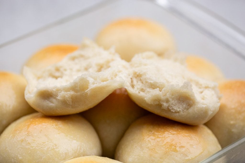 A close-up of several golden brown dinner rolls in a glass dish, with one roll split open to show its soft, fluffy interior.